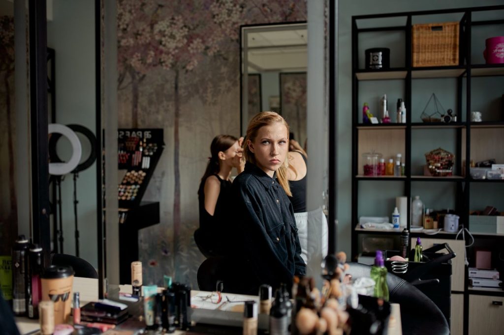 Side view of young attentive blond lady in black apparel looking in mirror near table with assorted cosmetic products in salon